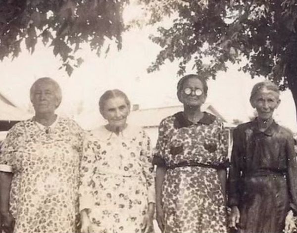 Four elderly women in floral dresses standing outdoors under a tree.
