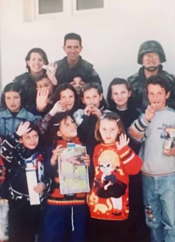 A group of children and soldiers posing together, some holding toys and waving at the camera.