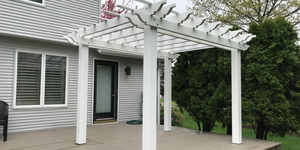 White pergola on a wooden deck attached to a house with gray siding.