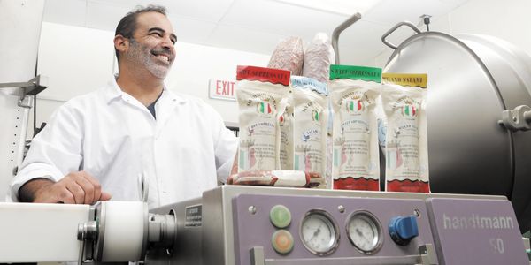Man in white coat smiles near sausage packaging machine.