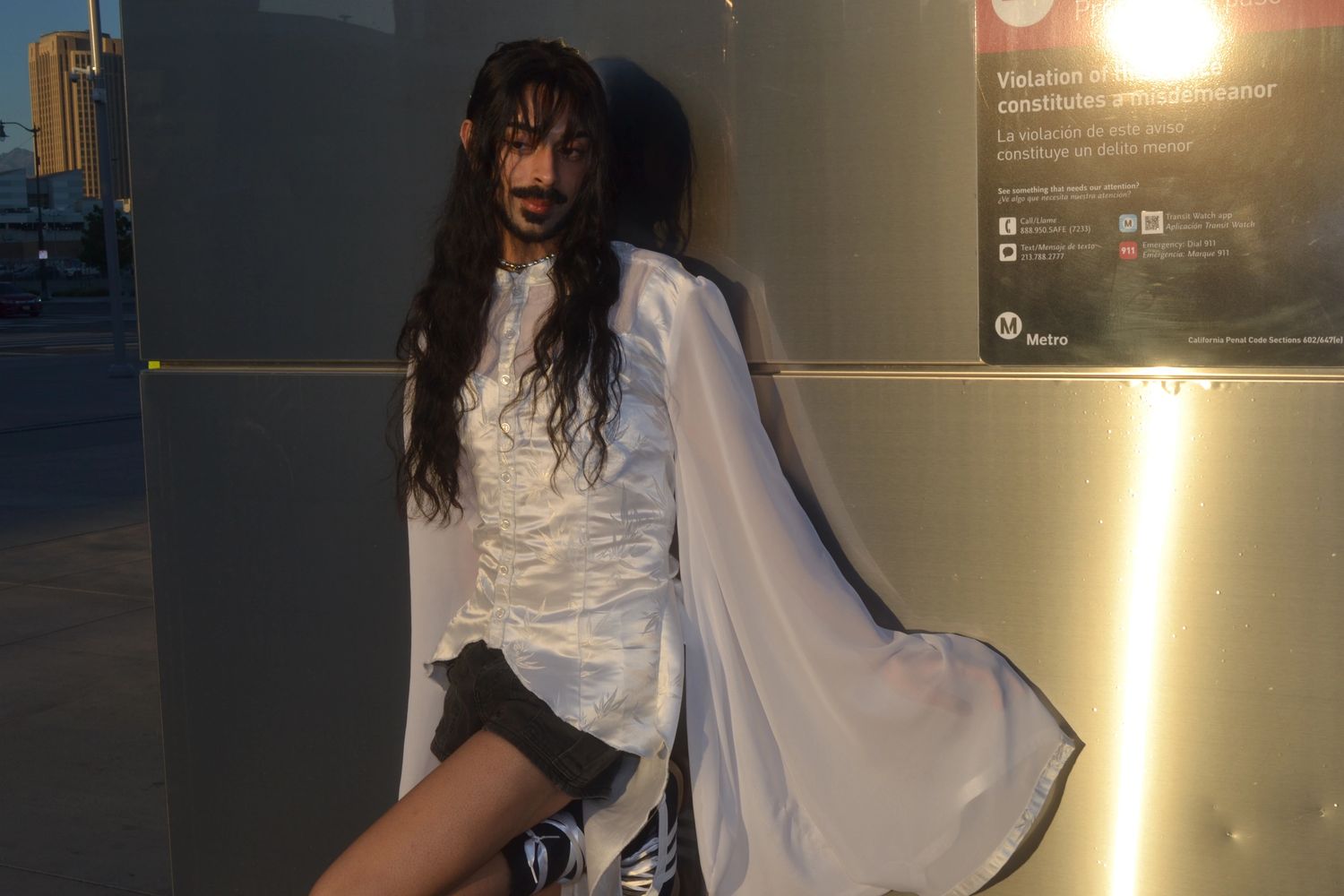 Person in flowing white blouse and black shorts leaning against a metallic wall at sunset.