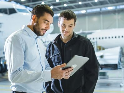 Two men reviewing information on a tablet inside an aircraft hangar.