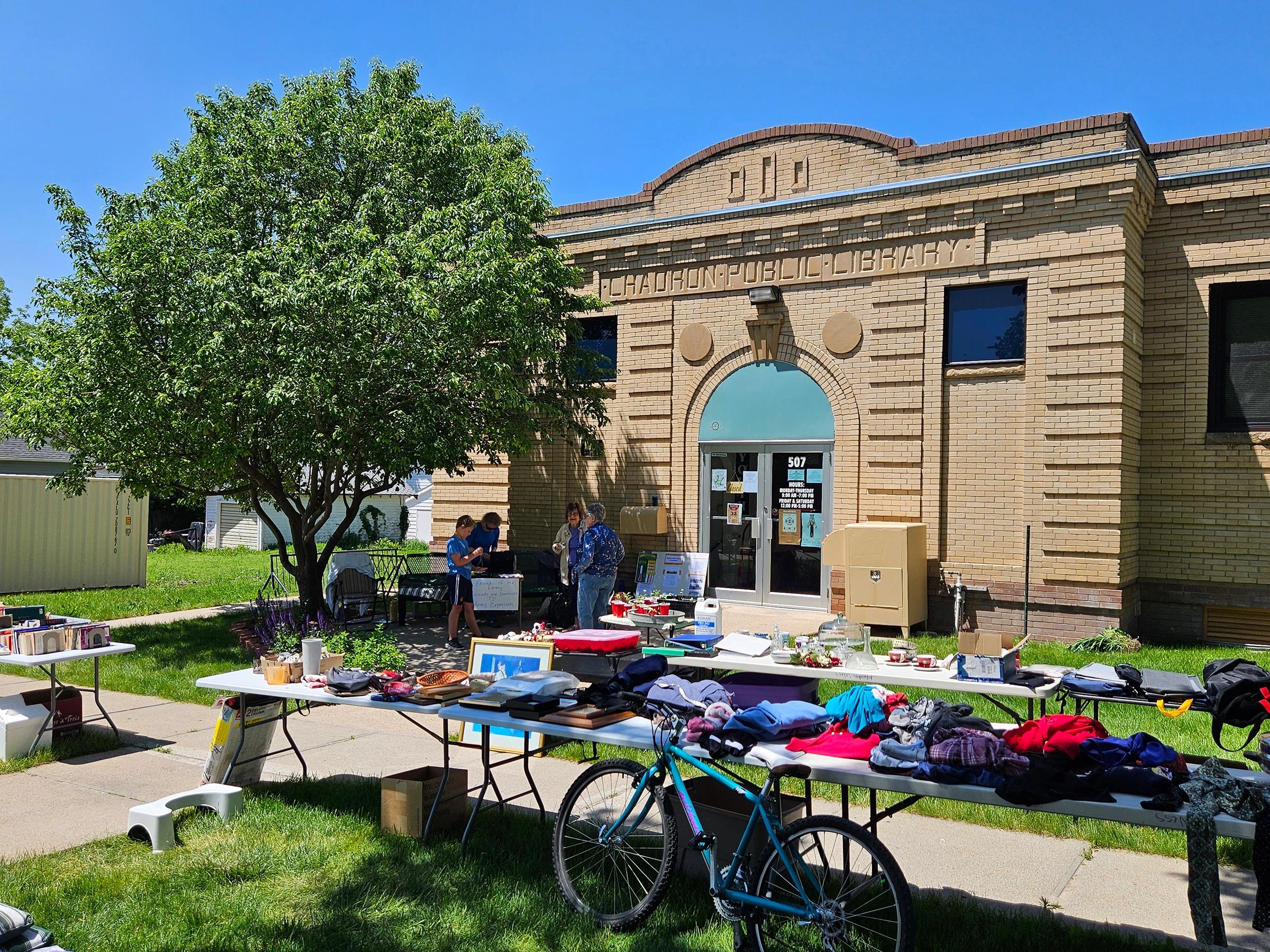 Shoppers browse garage sale items on tables in front of a brick library building. 