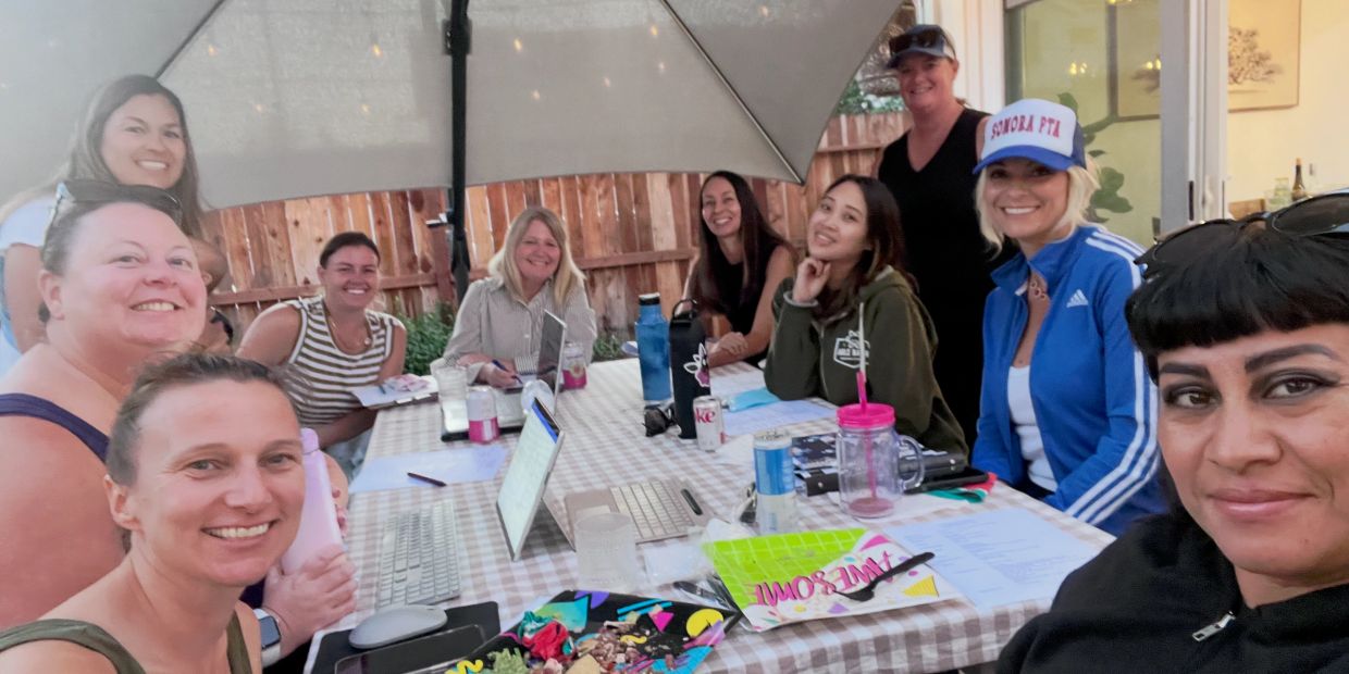 Group of smiling women gathered around an outdoor table with laptops and drinks.