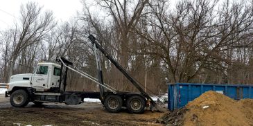 A white truck with a hydraulic system near a blue dumpster and dirt pile.