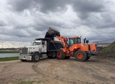 An orange loader dumping dirt into a white dump truck under a cloudy sky.