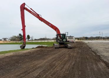 Excavator working on leveled dirt near a water body under a cloudy sky.