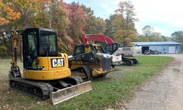 Construction vehicles lined up outdoors near a barn.