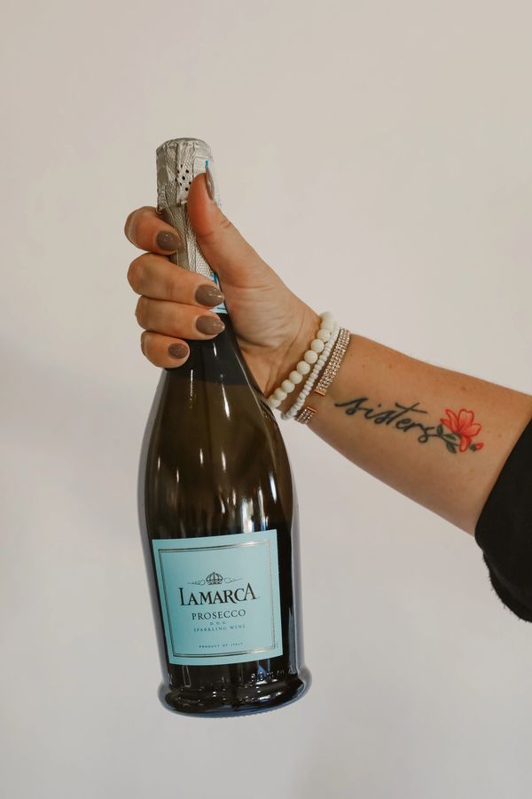 Hand holding a La Marca Prosecco bottle with a 'sisters' tattoo on the arm.
