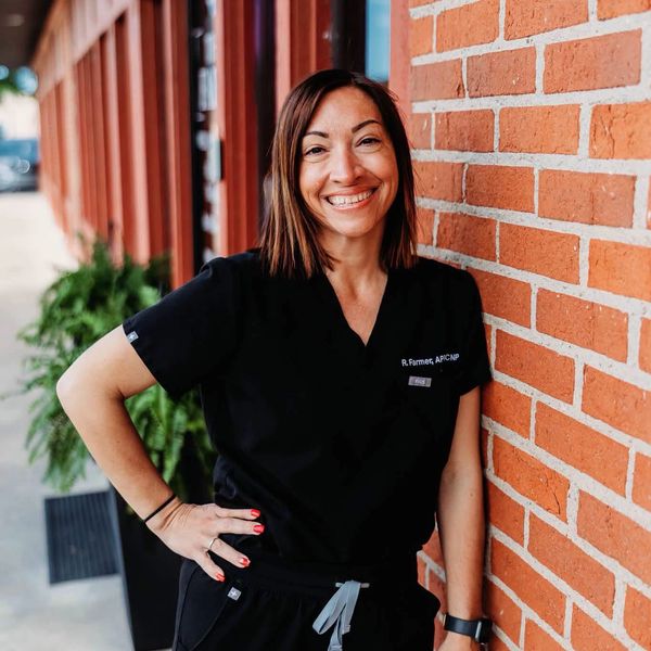 Smiling woman in black scrubs leaning against a brick wall.