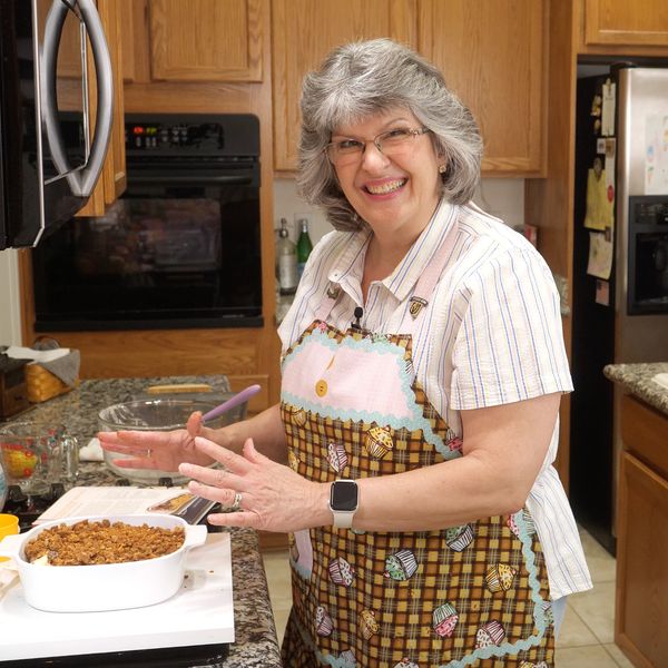 Smiling woman in a colorful apron presenting a baked dish in a kitchen.