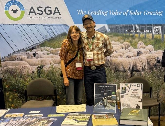 Two smiling people at the American Solar Grazing Association booth with informative materials and sheep backdrop.
