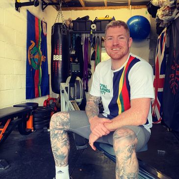 A man with tattoos sits on a bench in a home gym with boxing equipment.