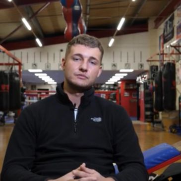 Man in black jacket sitting in a boxing gym with punching bags.