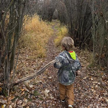 Child walking on a leaf-covered forest path holding a large stick.