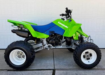 Bright green Kawasaki all-terrain quad bike with blue seat parked on pavement.