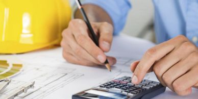 Person using calculator and pencil on architectural plans with a hard hat nearby.