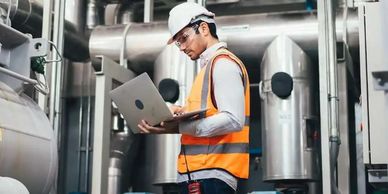 Engineer in safety gear working on laptop in an industrial facility.