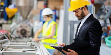 Engineer in a yellow helmet inspecting factory production line with clipboard.