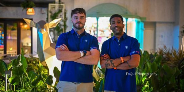 Two men in blue shirts standing with folded arms in a garden area.