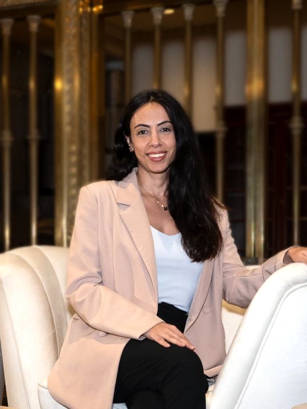 A woman with long hair smiles, sitting in an elegant chair wearing a beige blazer.