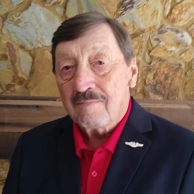 Elderly man in a blazer with a red shirt against a stone wall background.