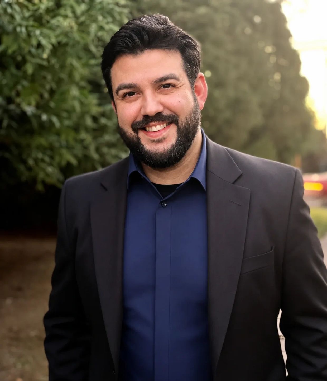 Man smiling outdoors in a blazer and blue shirt.