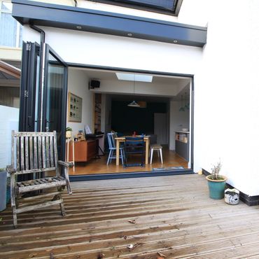 Open patio doors reveal a cozy dining area with wooden furniture and potted plants on a wooden deck.