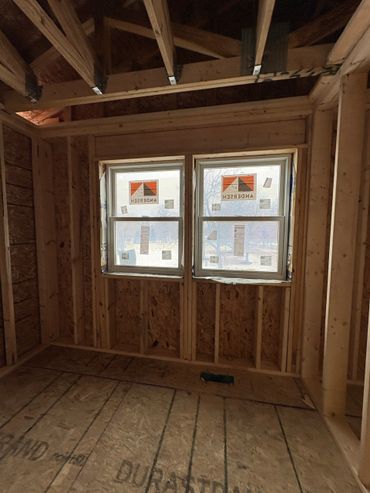 Interior view of a wooden-framed room under construction with two windows installed.