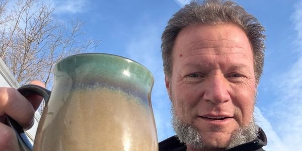 Smiling man holding a ceramic mug outdoors under a clear blue sky.