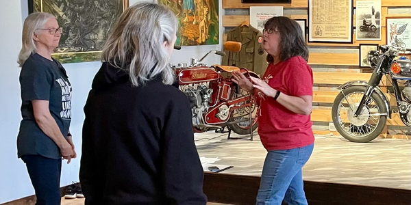 A woman explains vintage motorcycles to two listeners in a museum setting.