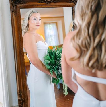 Bride in white dress holding a bouquet, looking into a mirror.