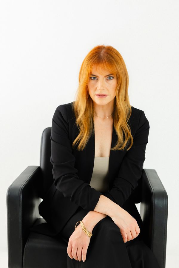 Confident red-haired woman in black suit sitting on a chair against a white background.