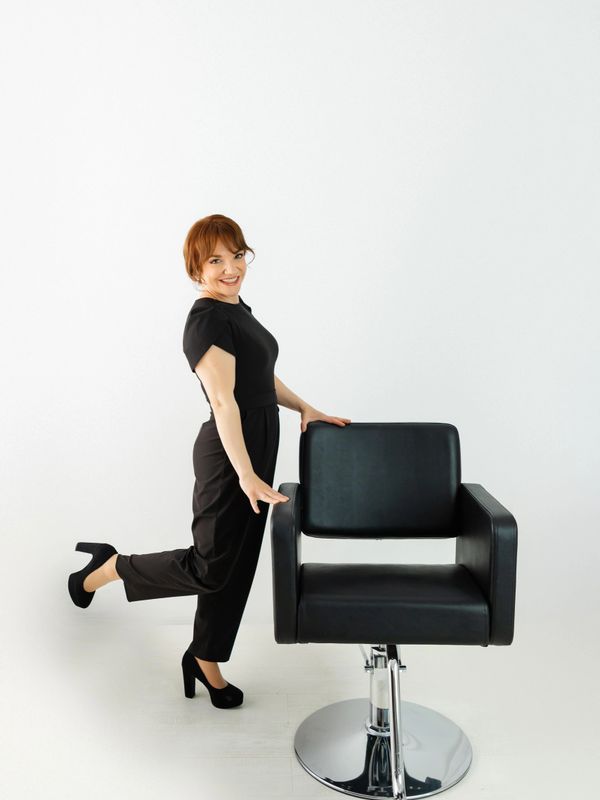 Woman in black outfit posing happily beside a black salon chair.