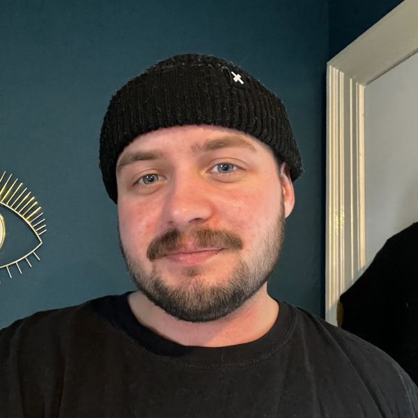 Alex wearing a black beanie and t-shirt, his eyes are blue and he has a stubble beard and moustache.