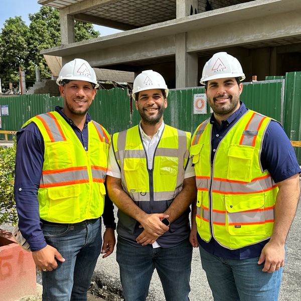 Three construction workers in safety vests and helmets smiling at a construction site.