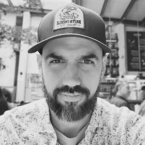 Portrait of a bearded man wearing a cap and floral shirt in a cafe.