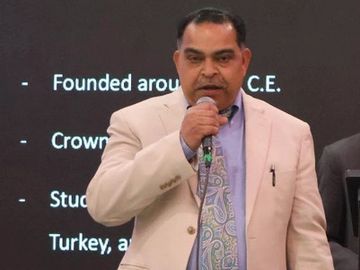 A man in a beige suit speaks into a microphone during a presentation.