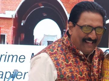 A man wearing glasses and a colorful vest smiles in front of a brick archway.