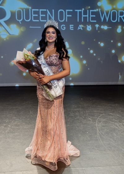 Beauty queen wearing crown and sash holding flowers on stage.