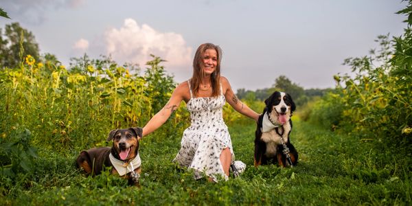 A woman in a white dress smiles with two dogs in a green field.