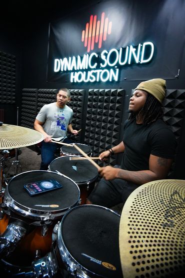 Two musicians play drums in a studio with Dynamic Sound Houston sign.