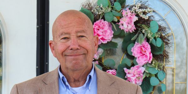 Smiling elderly man in a brown blazer stands in front of a door with a floral wreath.
