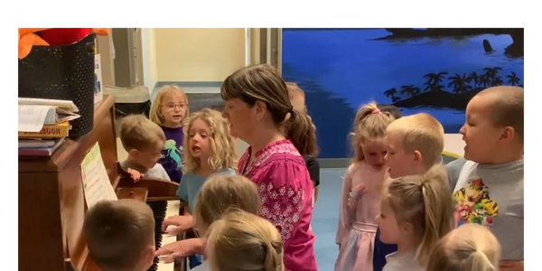 The director plays piano surrounded by children singing along in the early childhood education program that provides childcare services to children. 