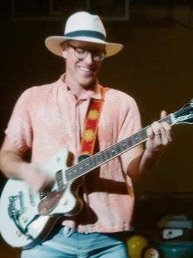 Man smiling and playing electric guitar wearing a hat and glasses.