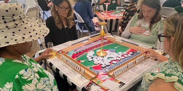 Four women playing Mahjong around a table indoors.