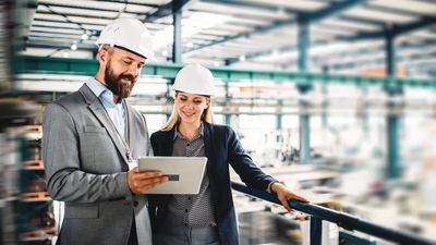 Two engineers in hard hats reviewing plans on a tablet in a factory.