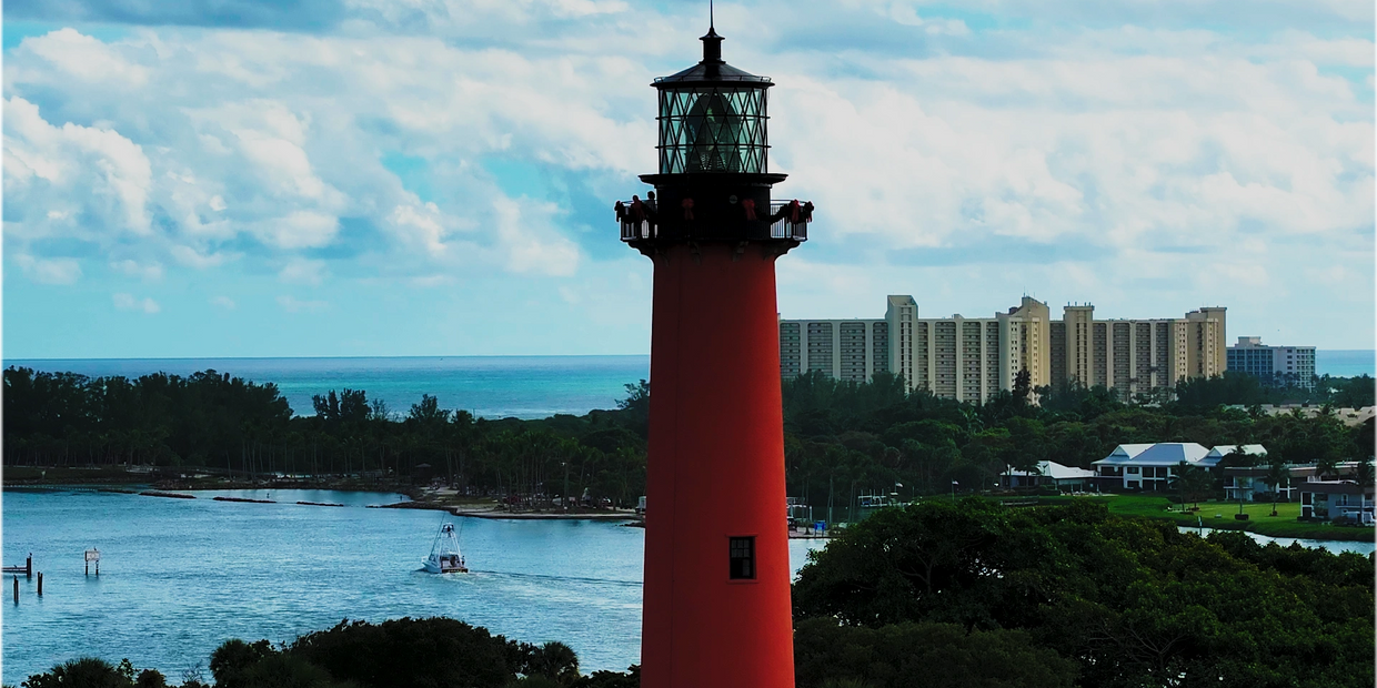 Tall red lighthouse overlooking water with buildings and greenery in the background.