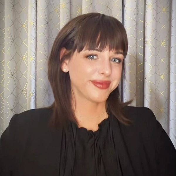 A woman with brown hair and bangs smiles warmly, wearing a black top against a patterned curtain backdrop.