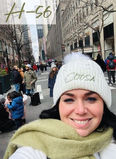 Woman smiling on a busy city street, wearing a white hat and green scarf.